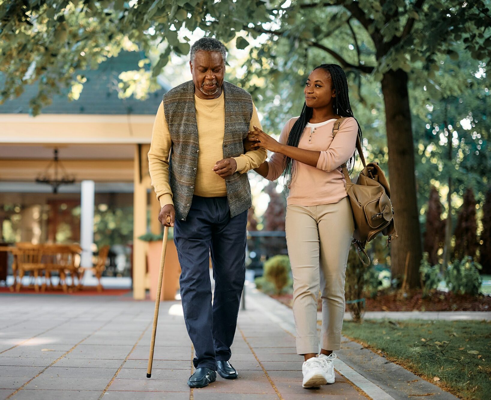 Black senior man with walking cane and his daughter a taking walk through park of nursing home.
